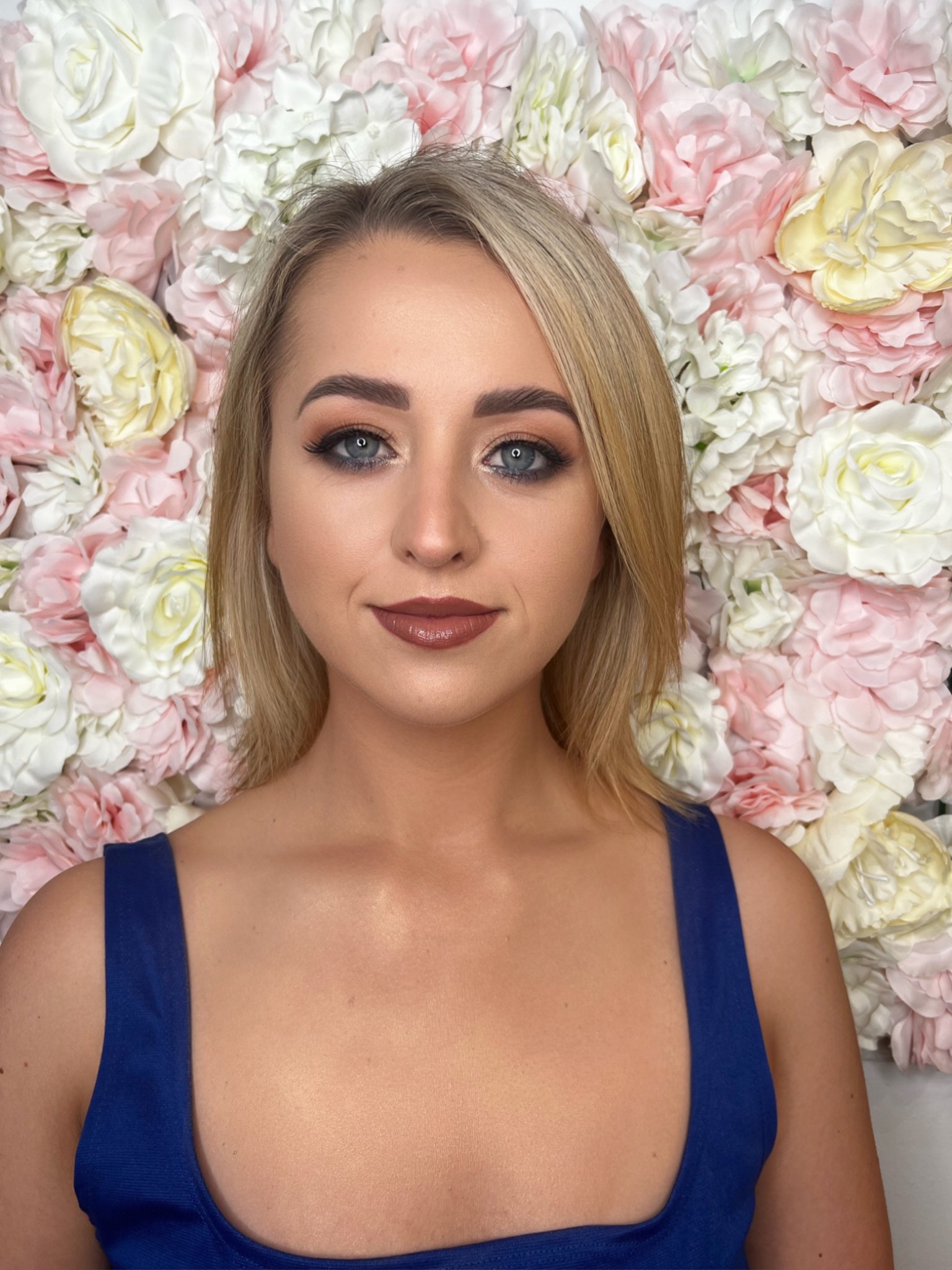 Portret blondynki z profesjonalnym makijażem oczu i ust w odcieniach brązu na tle ściany z białych i różowych kwiatów.