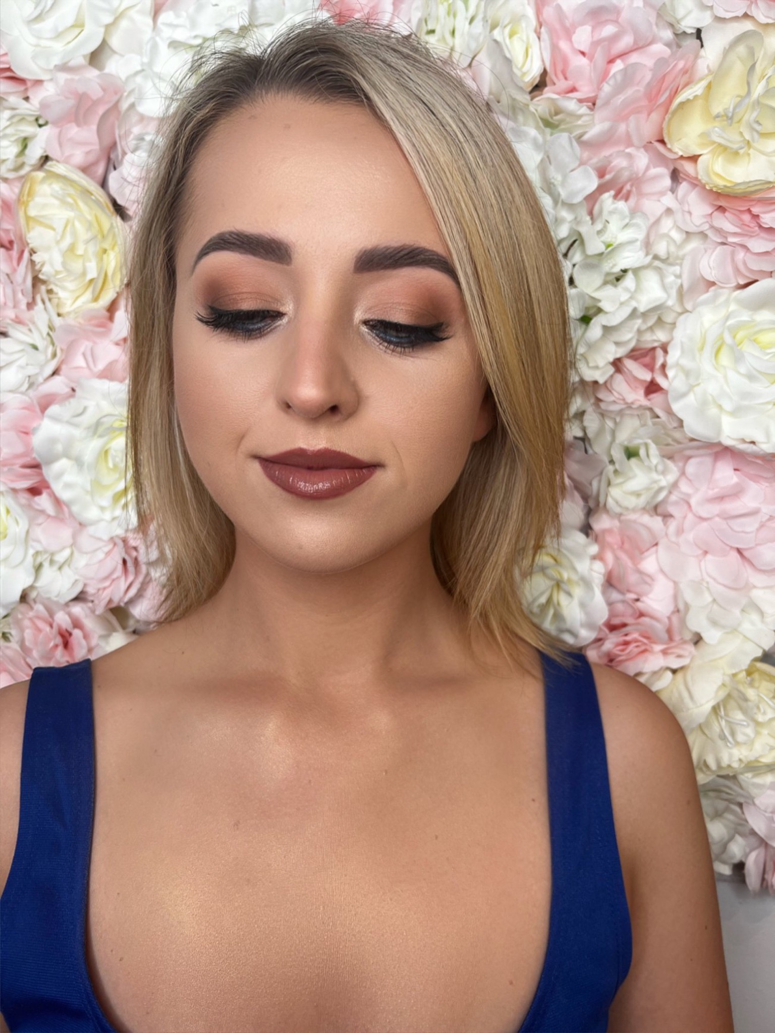 Portret kobiety z profesjonalnym makijażem w odcieniach brązu, z ustami w kolorze czekolady, na tle ściany z różowych i białych kwiatów.