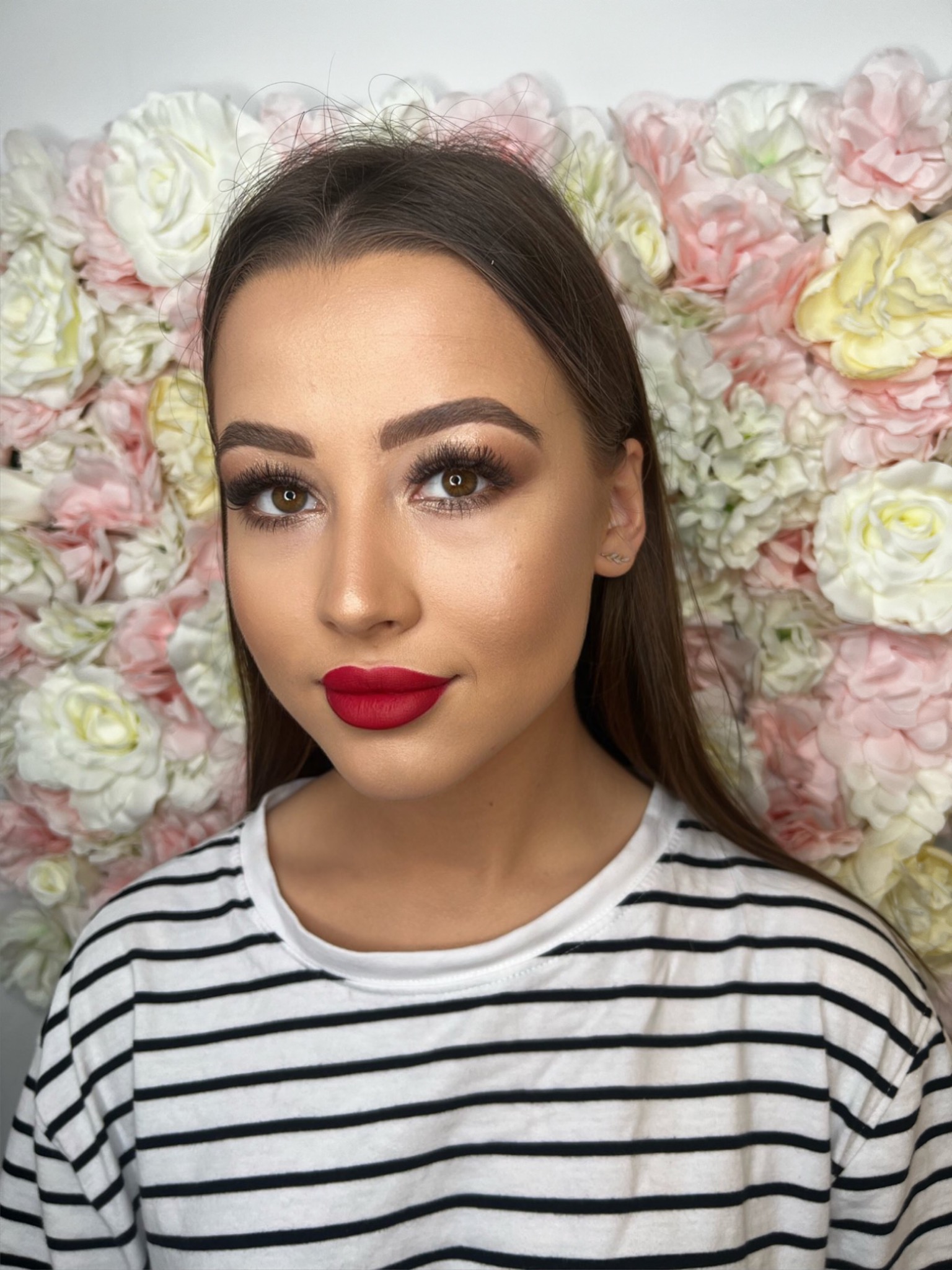 Portret kobiety z pełnym makijażem, w tym intensywnie czerwone usta i podkreślone oczy, na tle ściany z różowych i białych kwiatów.