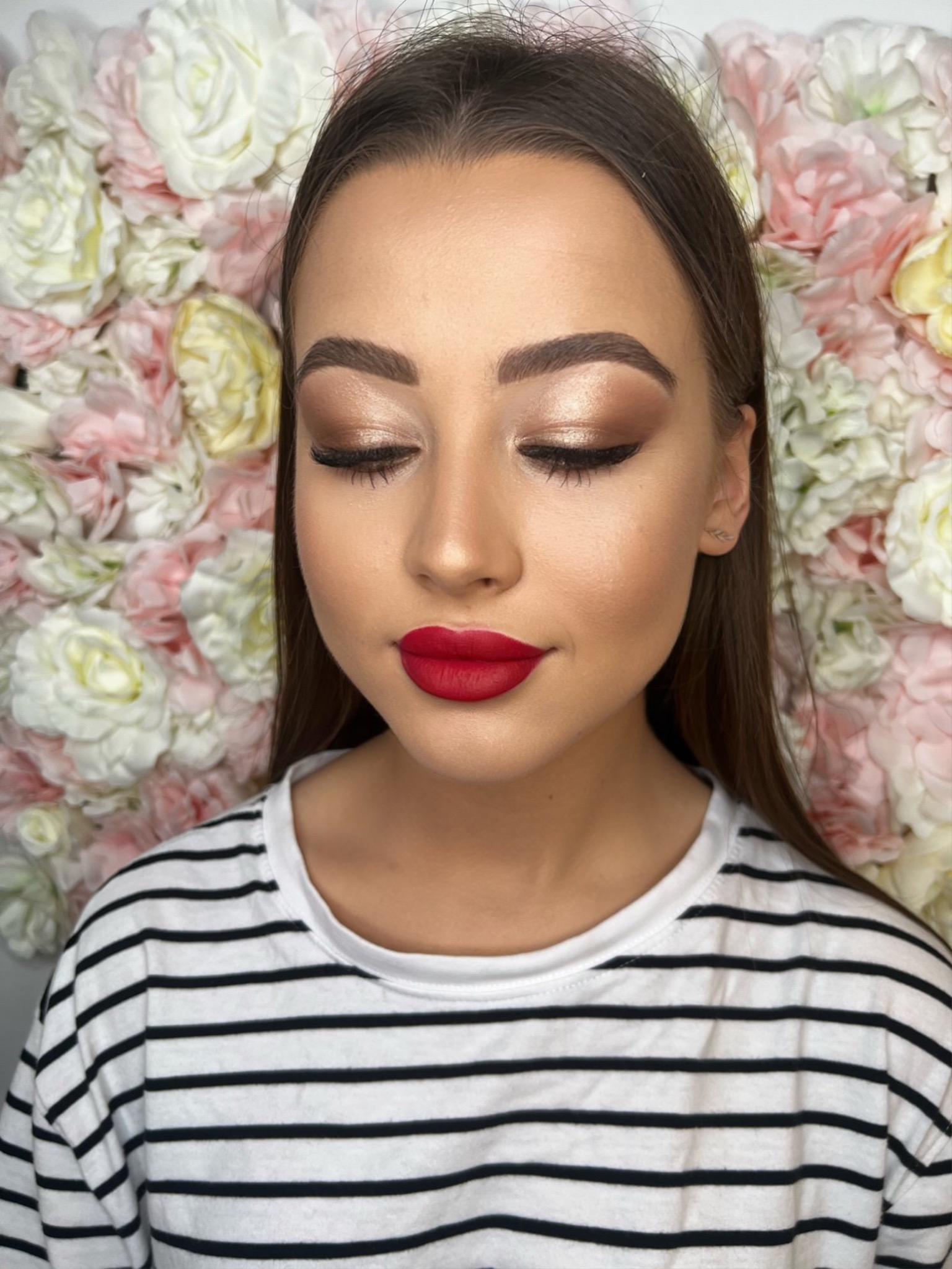 Portret kobiety z zamkniętymi oczami, prezentującej makijaż z błyszczącymi cieniami i intensywnie czerwoną szminką. W tle dekoracja z kwiatów.