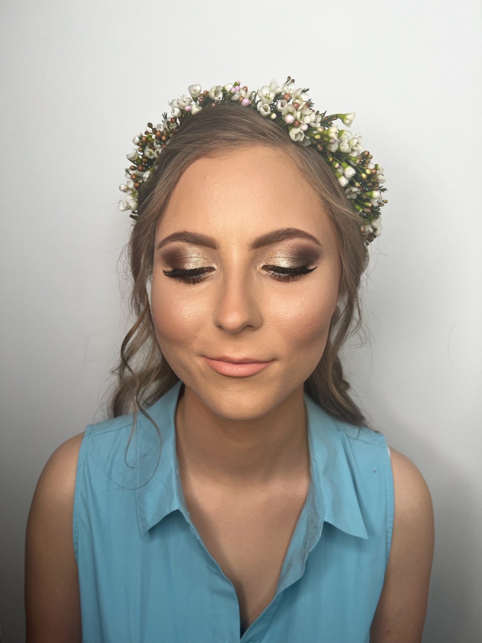 Portret kobiety z zamkniętymi oczami w pełnym makijażu ślubnym, wianek z drobnych kwiatów i perłowych koralików na głowie, delikatny uśmiech, złote cienie do powiek, długie rzęsy, konturowanie twarzy.