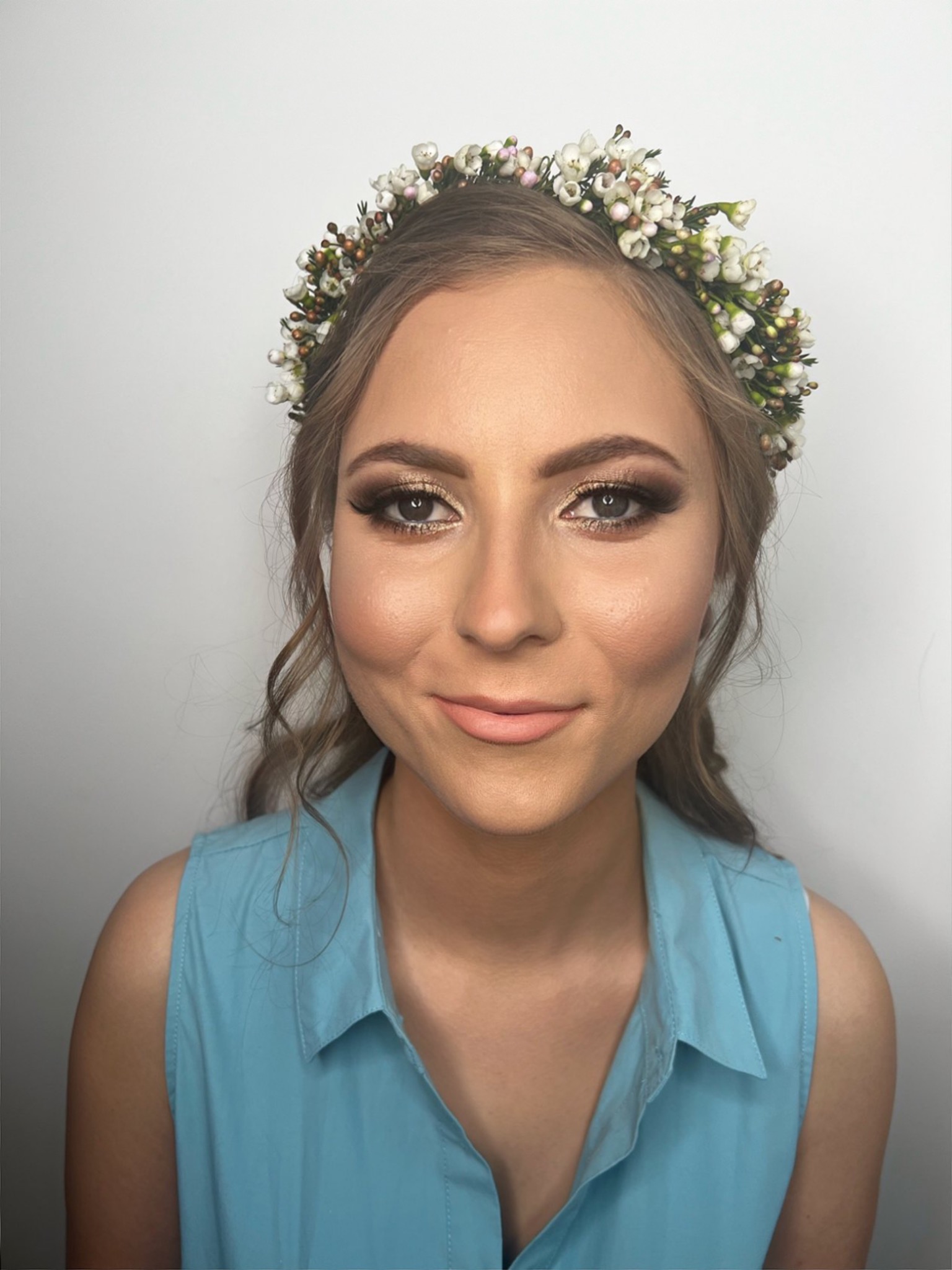 Portret kobiety z delikatnym makijażem oczu w odcieniach brązu i złotego oraz z wiankiem z białych kwiatów na głowie, ubrana w błękitną koszulę.