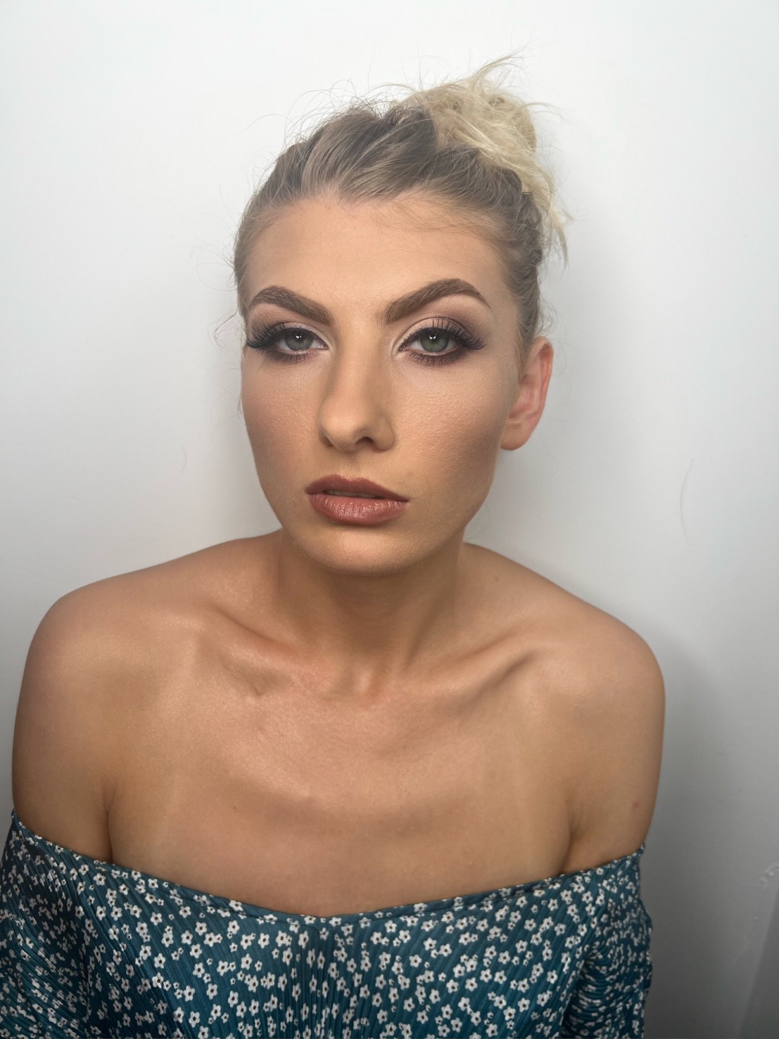 Portret kobiety z profesjonalnym makijażem: wyraziste oczy z długimi rzęsami, konturowana twarz i brązowe usta, włosy upięte w luźny kok na tle białej ściany.