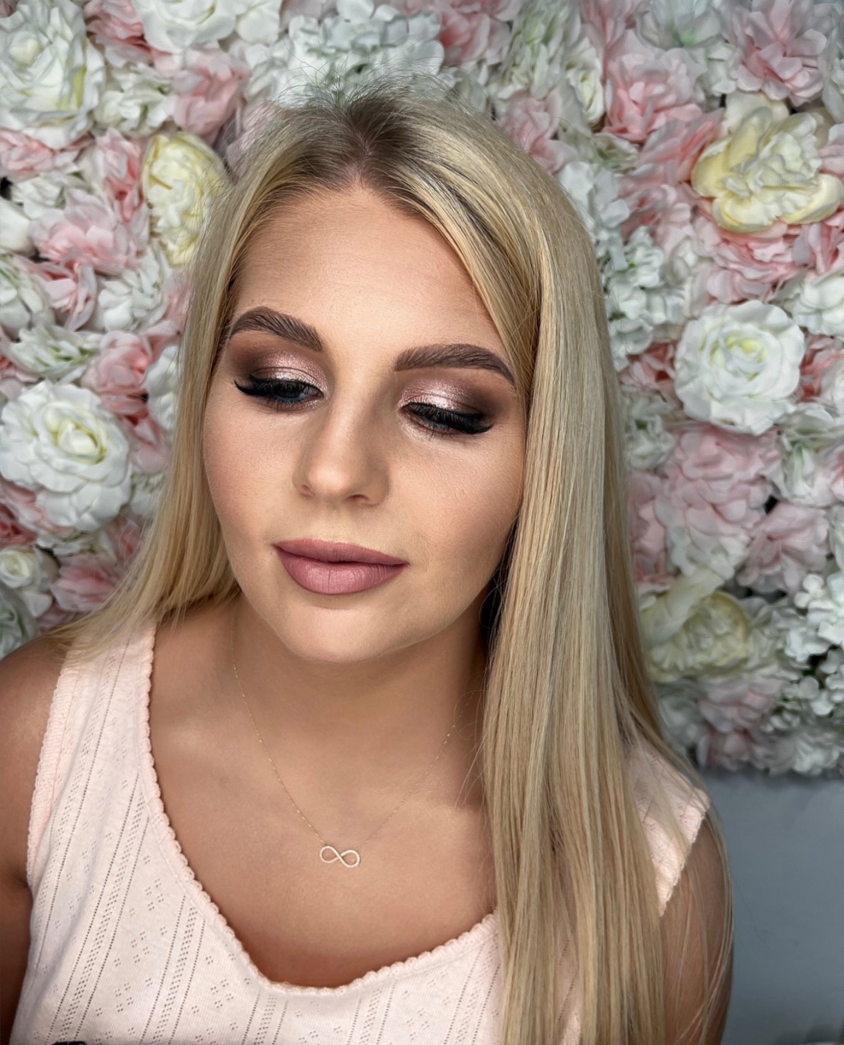 Portret blondynki z błyszczącym, brązowo-złotym makijażem oczu i matowymi, różowymi ustami, na tle ściany z różowych i białych kwiatów.