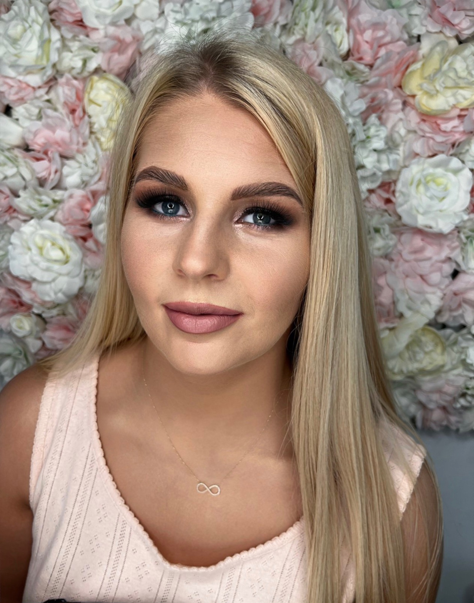 Portret blondynki z pełnym makijażem oczu i ust, na tle ściany z różowych i białych kwiatów.