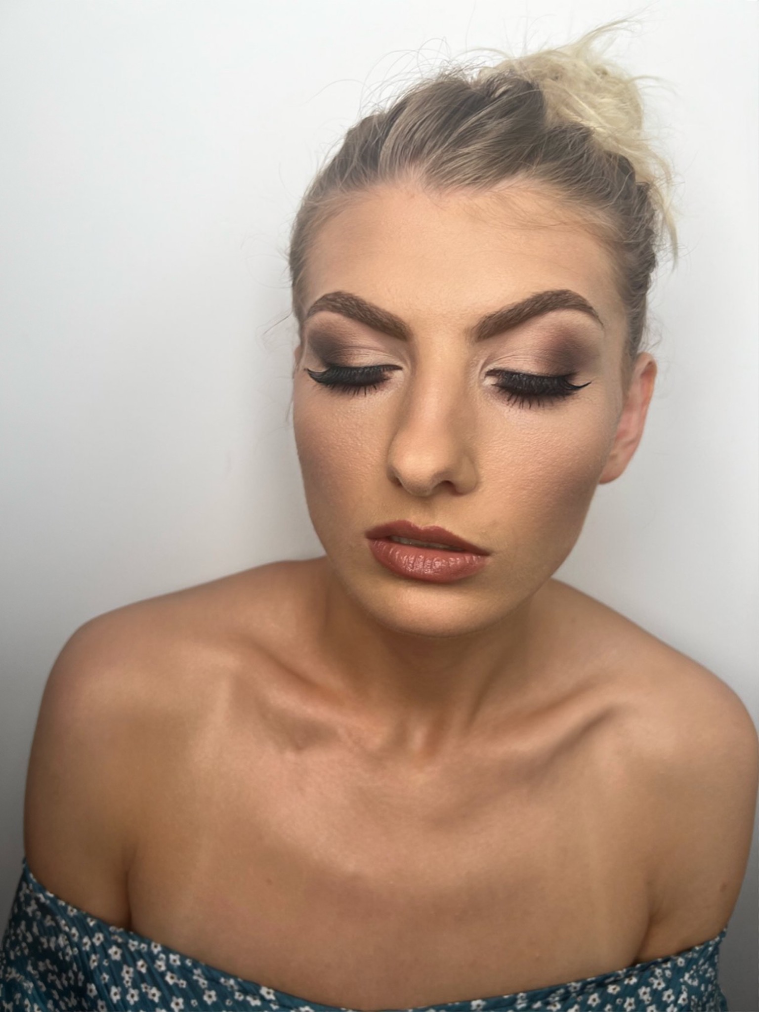 Portret kobiety z profesjonalnym makijażem oczu w odcieniach brązu i beżu, z mocno wytuszowanymi rzęsami i błyszczącymi ustami w kolorze nude, włosy upięte w luźny kok na czubku głowy, widoczny...