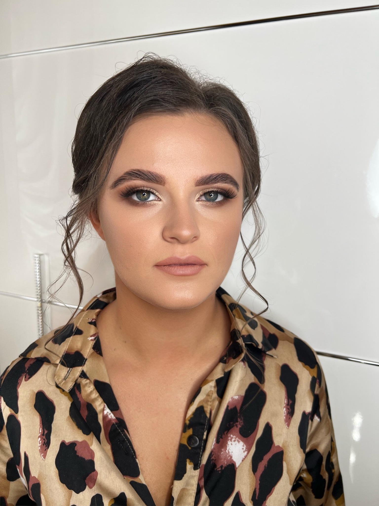 Portret kobiety z profesjonalnym makijażem oczu w odcieniach brązu i beżu, podkreślone brwi, włosy upięte z luźnymi kosmykami, ubrana w koszulę w panterkę na tle białej ściany.