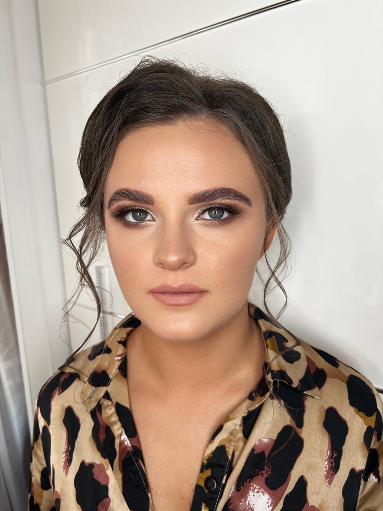 Portret kobiety z profesjonalnym makijażem: podkreślone brwi, złoty cień na powiekach, wydłużone rzęsy i delikatny makijaż ust, ubrana w koszulę w panterkę.