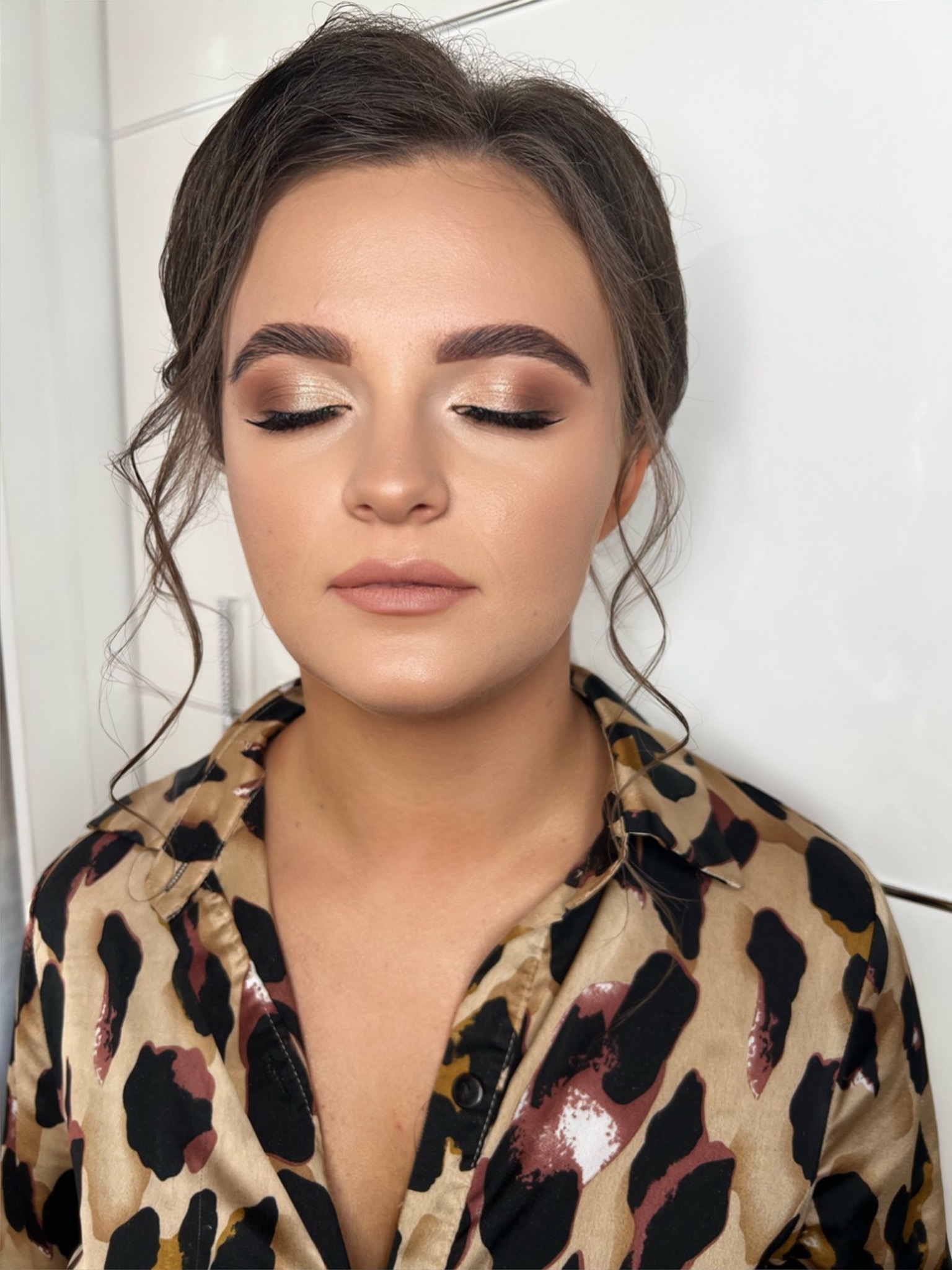 Portret kobiety z zamkniętymi oczami, prezentujący delikatny, złoty makijaż oczu z czarną kreską, naturalne brwi i subtelne podkreślenie ust, ubrana w koszulę w panterkę.