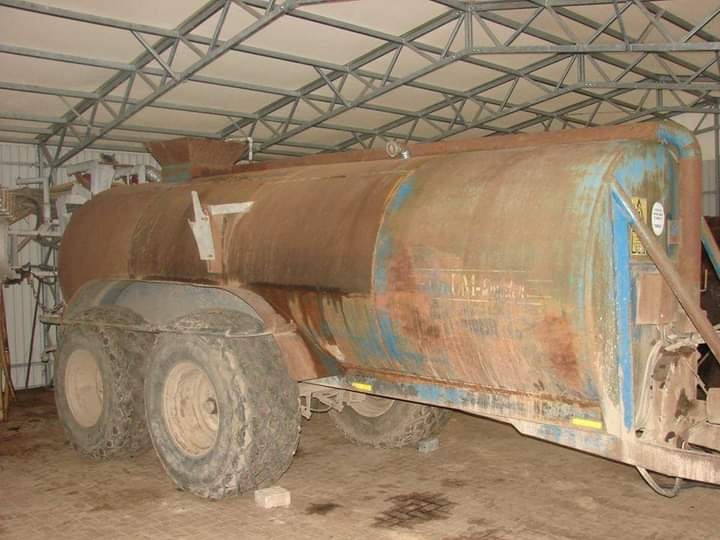 Stara przyczepa rolnicza z metalowym zbiornikiem pokrytym rdzą, stojąca wewnątrz hali z metalową konstrukcją dachu.