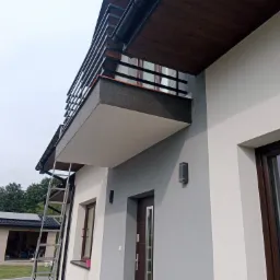 Elewacja domu z balkonem, widoczna drabina oparta o ścianę, budynek wykończony tynkiem w dwóch odcieniach szarości, z brązowymi elementami wykończeniowymi.