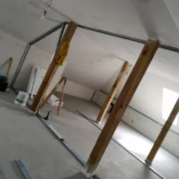 Poddasze w trakcie ocieplania z widoczną konstrukcją metalową, drewnianymi belkami podtrzymującymi i wiszącą żarówką na kablu.