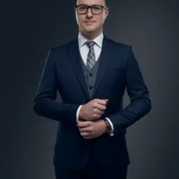 Portret mężczyzny w okularach, eleganckim granatowym garniturze, szarej kamizelce i wzorzystym krawacie, z zegarkiem na ręku, pozującego na ciemnoszarym tle.