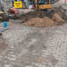 Żółta koparka CAT 302.7 na budowie, wykop zabezpieczony siatką, tablica ostrzegawcza 'Uwaga! Głębokie wykopy', w tle budynek w rusztowaniach i stos rur.