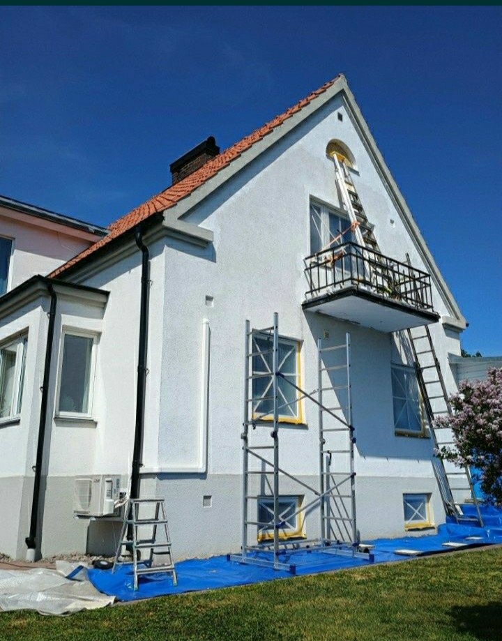 Biały dom z balkonem w trakcie odświeżania elewacji, okna zabezpieczone taśmą, rusztowania i drabiny rozstawione na niebieskiej folii ochronnej.