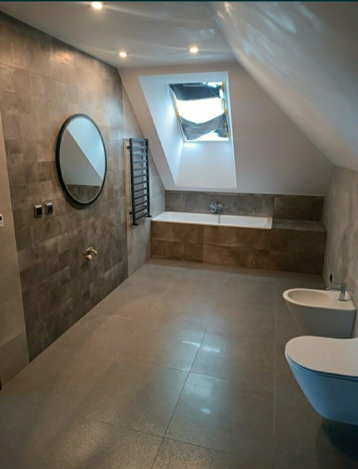 Wykończona łazienka na poddaszu z oknem dachowym, szarą glazurą, owalnym lustrem i grzejnikiem drabinkowym.