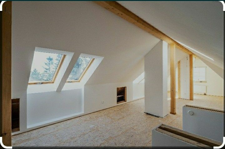 Poddasze w trakcie wykańczania: białe ściany, dwa okna dachowe z widokiem na zimowe drzewa, surowe drewniane belki stropowe i kolumny, podłoga z płyt OSB.