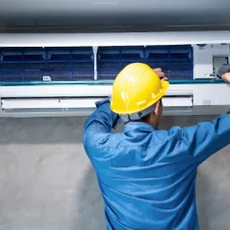 Mężczyzna w niebieskiej koszuli i żółtym kasku serwisuje klimatyzator ścienny, z otwartą obudową i widocznymi filtrami, używając specjalistycznego narzędzia do czyszczenia.