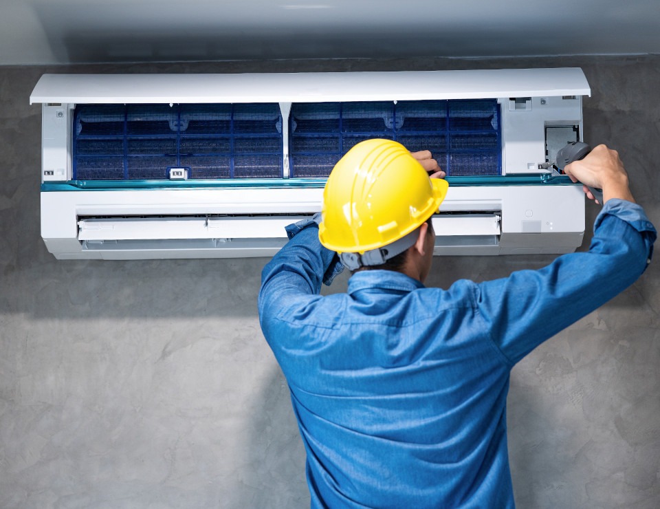 Mężczyzna w niebieskiej koszuli i żółtym kasku serwisuje klimatyzator ścienny, z otwartą obudową i widocznymi filtrami, używając specjalistycznego narzędzia do czyszczenia.