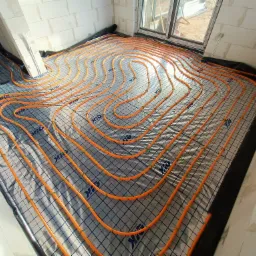 Pomarańczowe rury ogrzewania podłogowego ułożone spiralnie na czarnej folii z nadrukiem, przymocowane zielonymi spinkami, widoczne ściany z betonu komórkowego i okno.