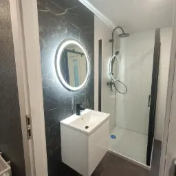 Nowoczesna łazienka z kabiną prysznicową z czarnymi ramami, białą szafką pod umywalką z czarną baterią i okrągłym lustrem LED na ścianie z imitacją czarnego marmuru.