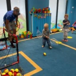 Nauczyciel prowadzi zajęcia sportowe z małymi dziećmi w jasnym, kolorowym pomieszczeniu. Dzieci trzymają rakiety i stoją na tle regału z zabawkami.