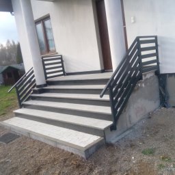 OgrodzeniaBS - Zewnętrzne schody z szarej kostki brukowej prowadzące do wejścia domu, z czarną metalową balustradą. Widoczne niedokończone ocieplenie ściany. Otoczenie to ziemia z kamieniami.