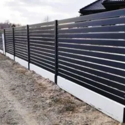 Długi, poziomy, czarny płot z poziomych desek z białymi podmurówkami, widziany z boku, z otwartą furtką i żwirową drogą w tle.