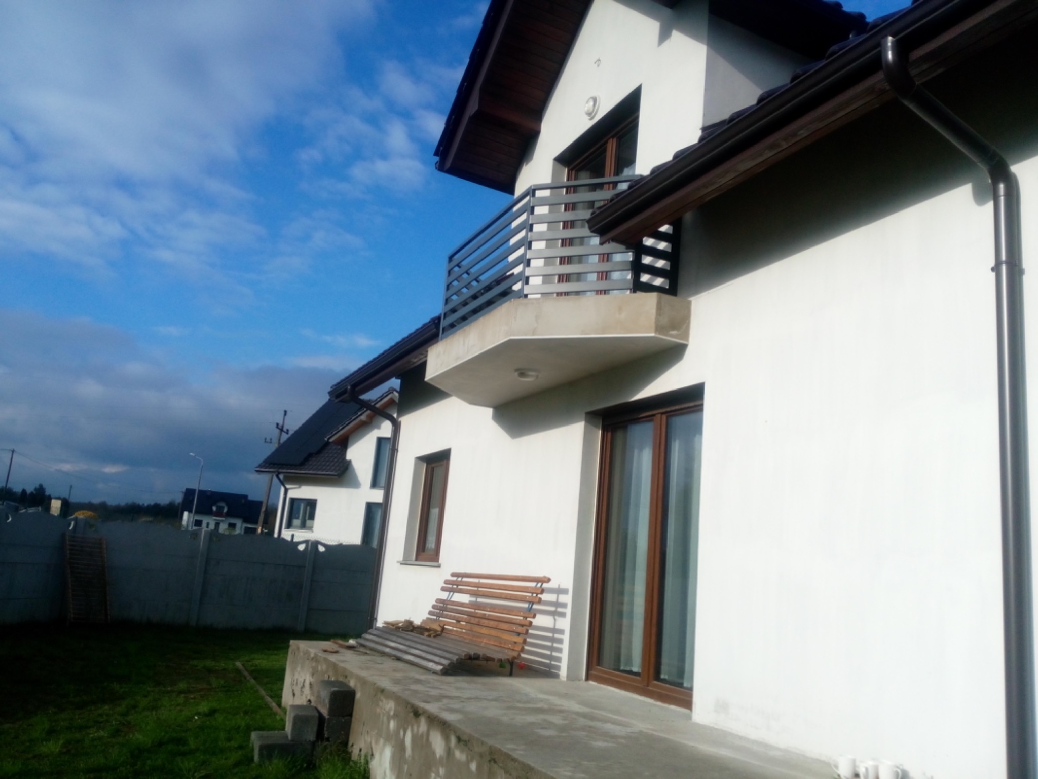 Nowoczesny dom z balkonem z metalową balustradą, widok z boku na tle pochmurnego nieba, z widocznym fragmentem betonowego ogrodzenia i drewnianej ławki na tarasie.