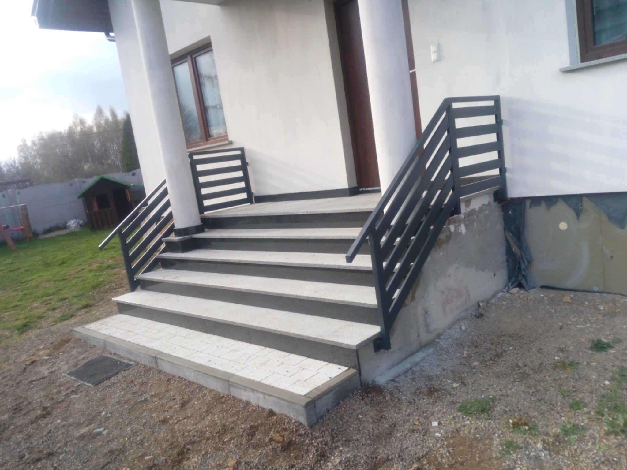Zewnętrzne schody z szarej kostki brukowej prowadzące do wejścia domu, z czarną metalową balustradą. Widoczne niedokończone ocieplenie ściany. Otoczenie to ziemia z kamieniami.