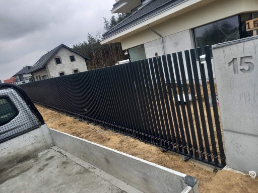 Nowoczesne, czarne ogrodzenie panelowe z pionowych profili, zakończone betonowym słupkiem z numerem domu '15', widoczne z perspektywy skrzyni ładunkowej samochodu dostawczego, w tle domy...