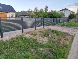 Nowoczesne, poziome ogrodzenie panelowe w kolorze grafitowym na betonowej podmurówce, widoczne słupki i brama w tle domy jednorodzinne i zieleń.