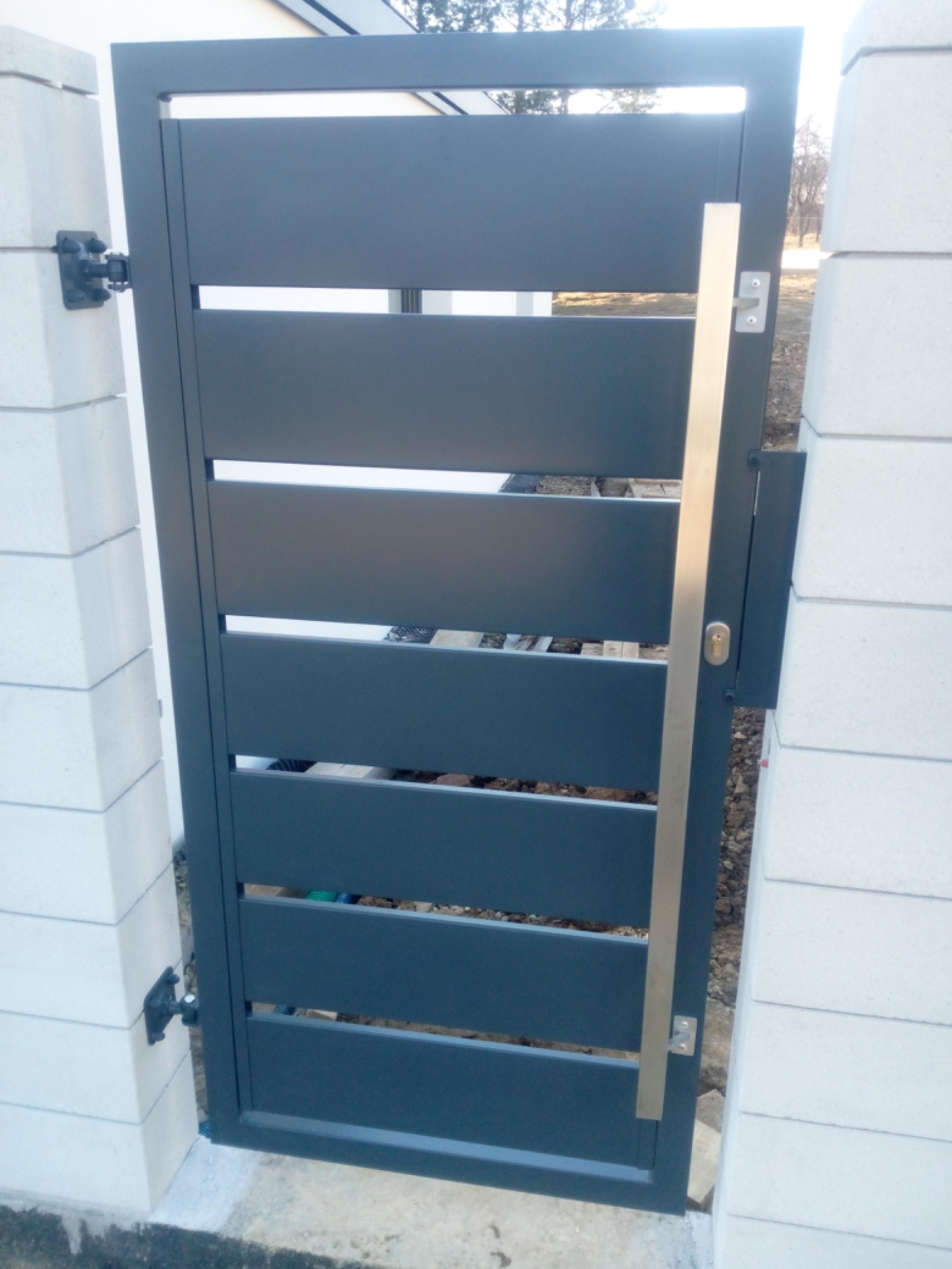 Nowoczesna, grafitowa furtka z poziomymi panelami, zamontowana między białymi, betonowymi słupkami, z widocznym srebrnym uchwytem i zamkiem.