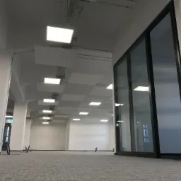 Nowoczesne biuro z podwieszanymi panelami LED, widoczne koryta kablowe pod sufitem, duże szklane drzwi przesuwne z czarnymi ramami.