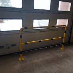 Żółta metalowa bariera ochronna z czarnymi pasami ostrzegawczymi, zamontowana wewnątrz hali przemysłowej przed bramą segmentową z oknami.