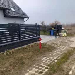 Nowoczesna, pozioma, szara konstrukcja oddzielająca posesję od chodnika z kostki ażurowej, na tle domu z szarą dachówką i czerwoną hydrantu.