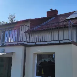 Dom z balkonem zabezpieczonym czarną, metalową balustradą, widoczne panele słoneczne na dachu i fragment ogrodu w słoneczny dzień.