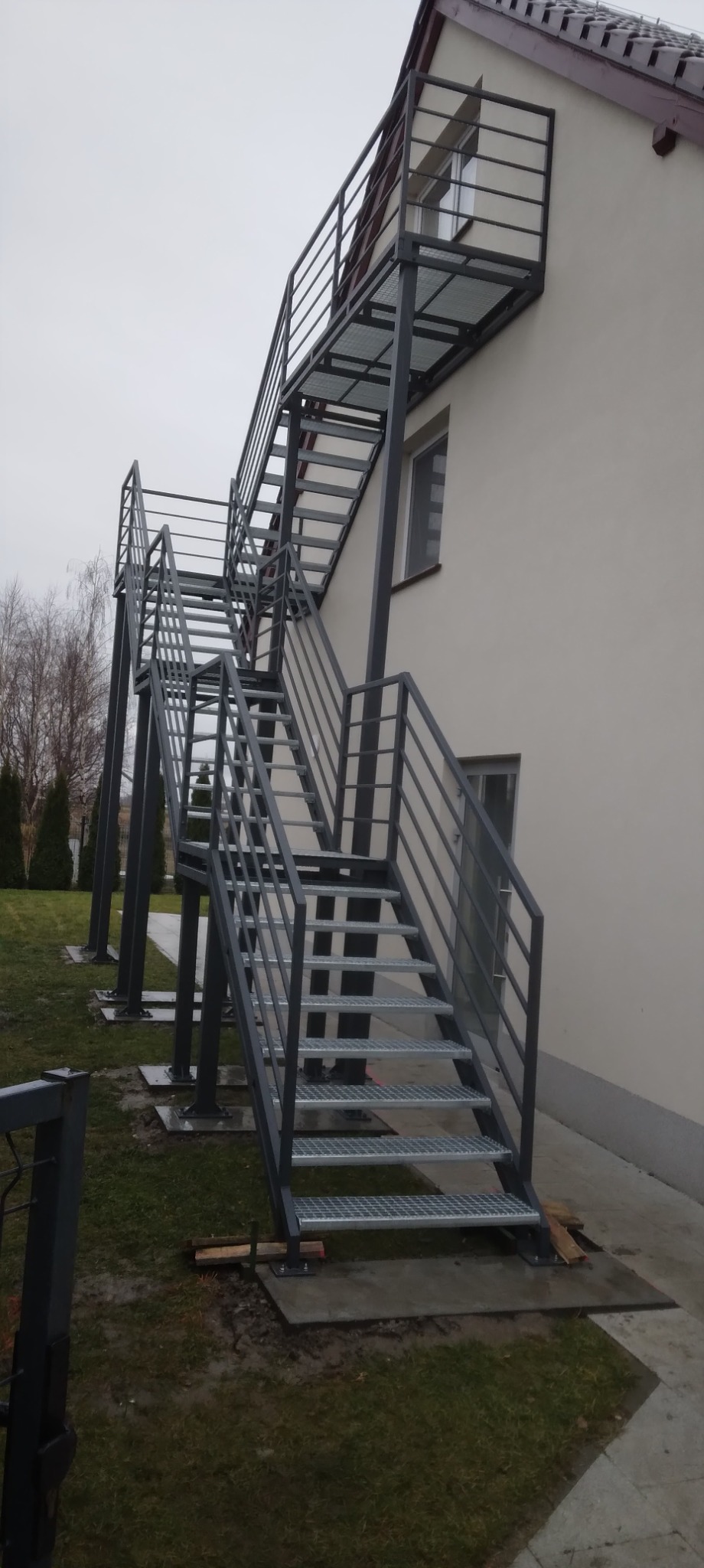 Zewnętrzne schody metalowe z platformą, prowadzące do okna na piętrze, z ażurowymi stopniami i prostymi balustradami, zamontowane przy budynku z jasną elewacją.