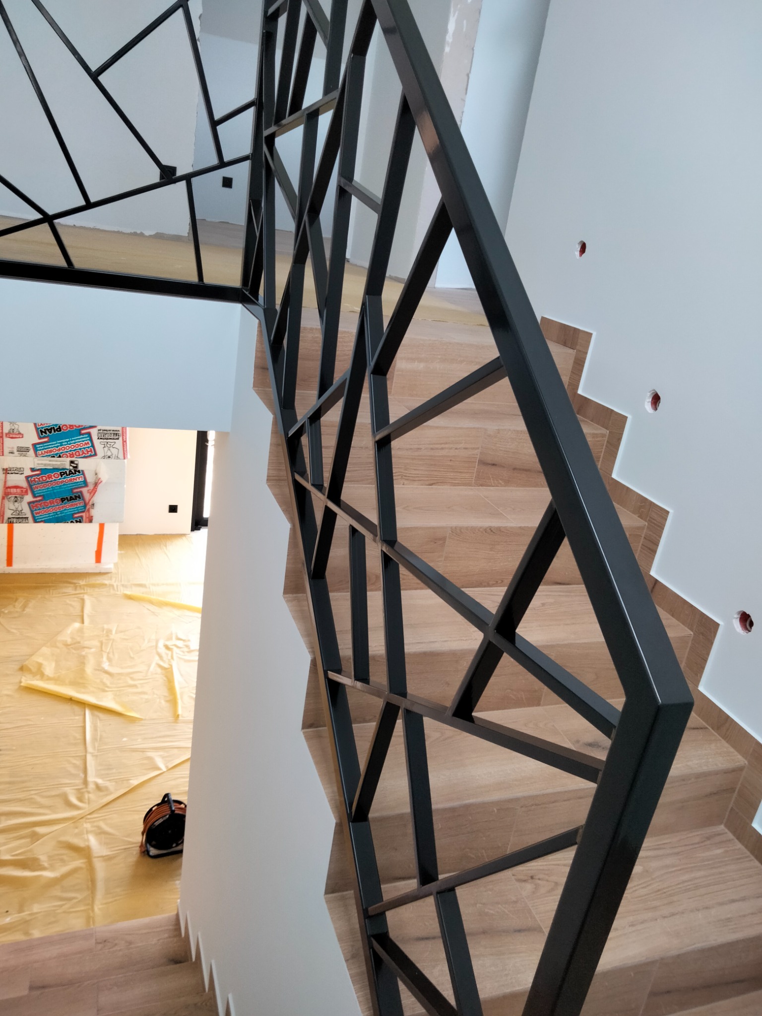 Schody z drewnopodobnymi płytkami i czarną, geometryczną balustradą. Widok z góry, w tle folia ochronna na podłodze i materiały budowlane.