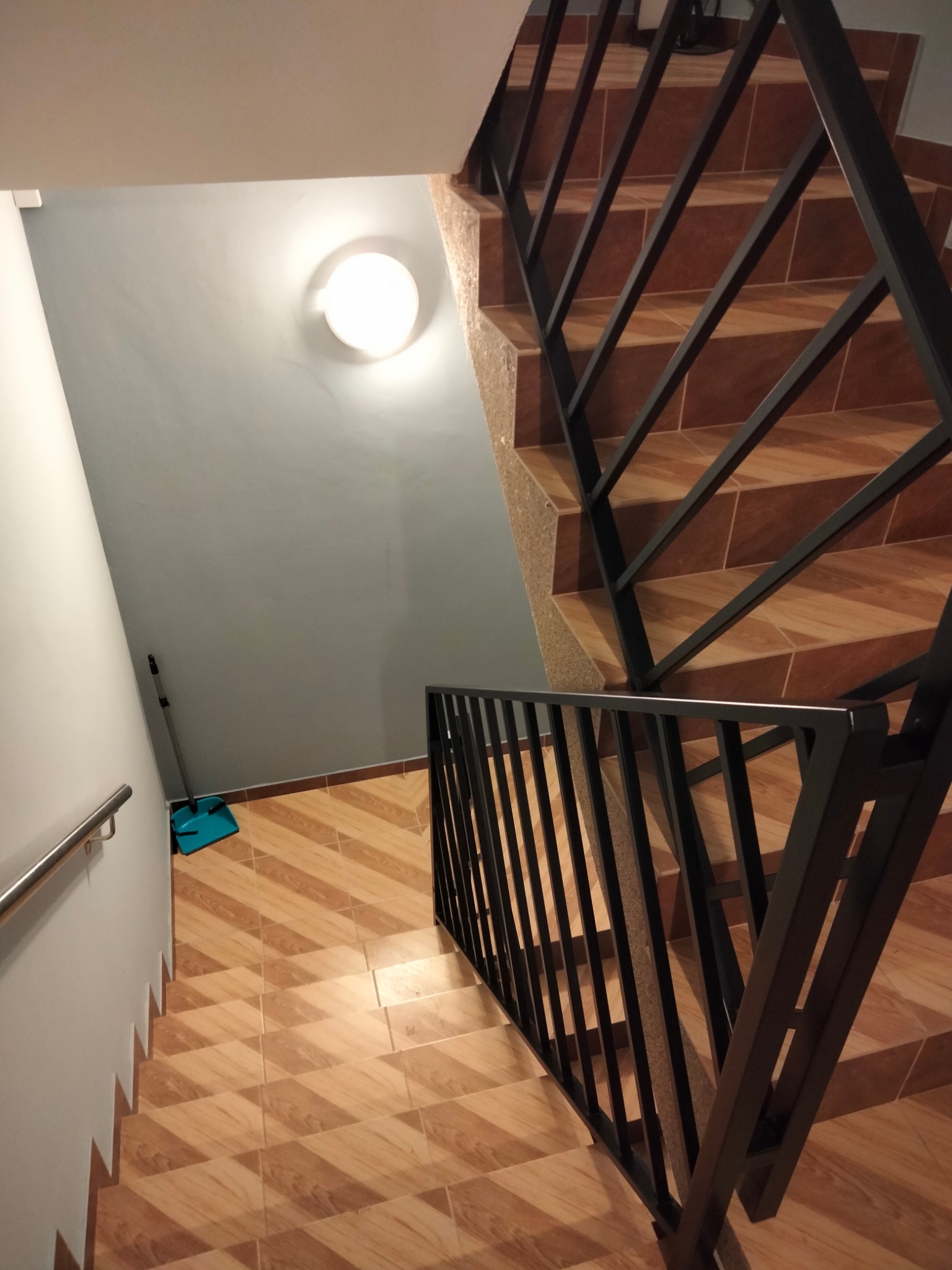 Wnętrze z brązowymi schodami wyłożonymi płytkami i czarną, metalową balustradą o geometrycznym wzorze. Widoczne oświetlenie i fragment ściany z poręczą.