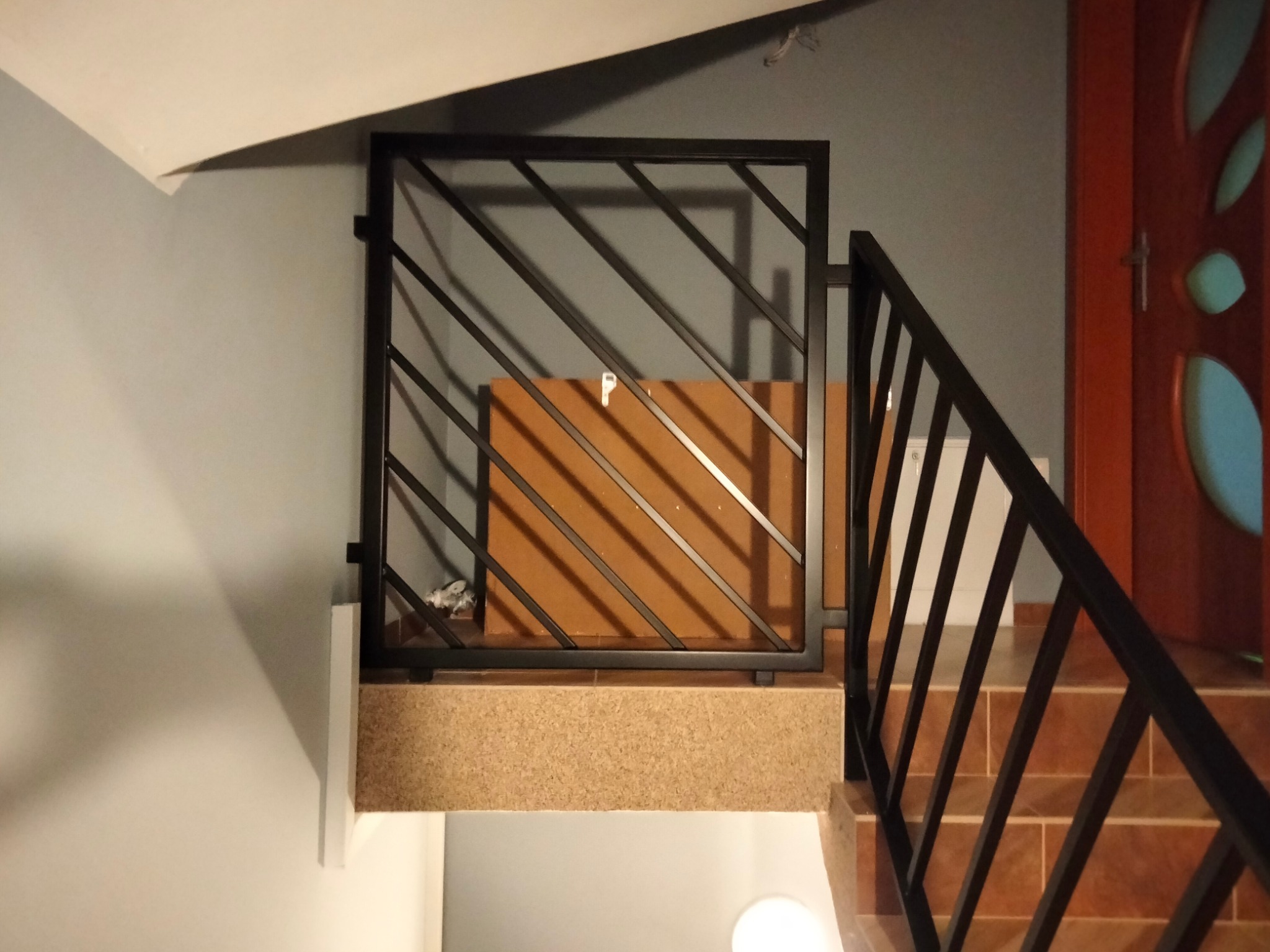 Nowoczesna, czarna barierka schodowa z ukośnymi prętami, Jabłówko. Metalowa konstrukcja zabezpiecza schody, widoczne brązowe płytki i fragment drzwi.