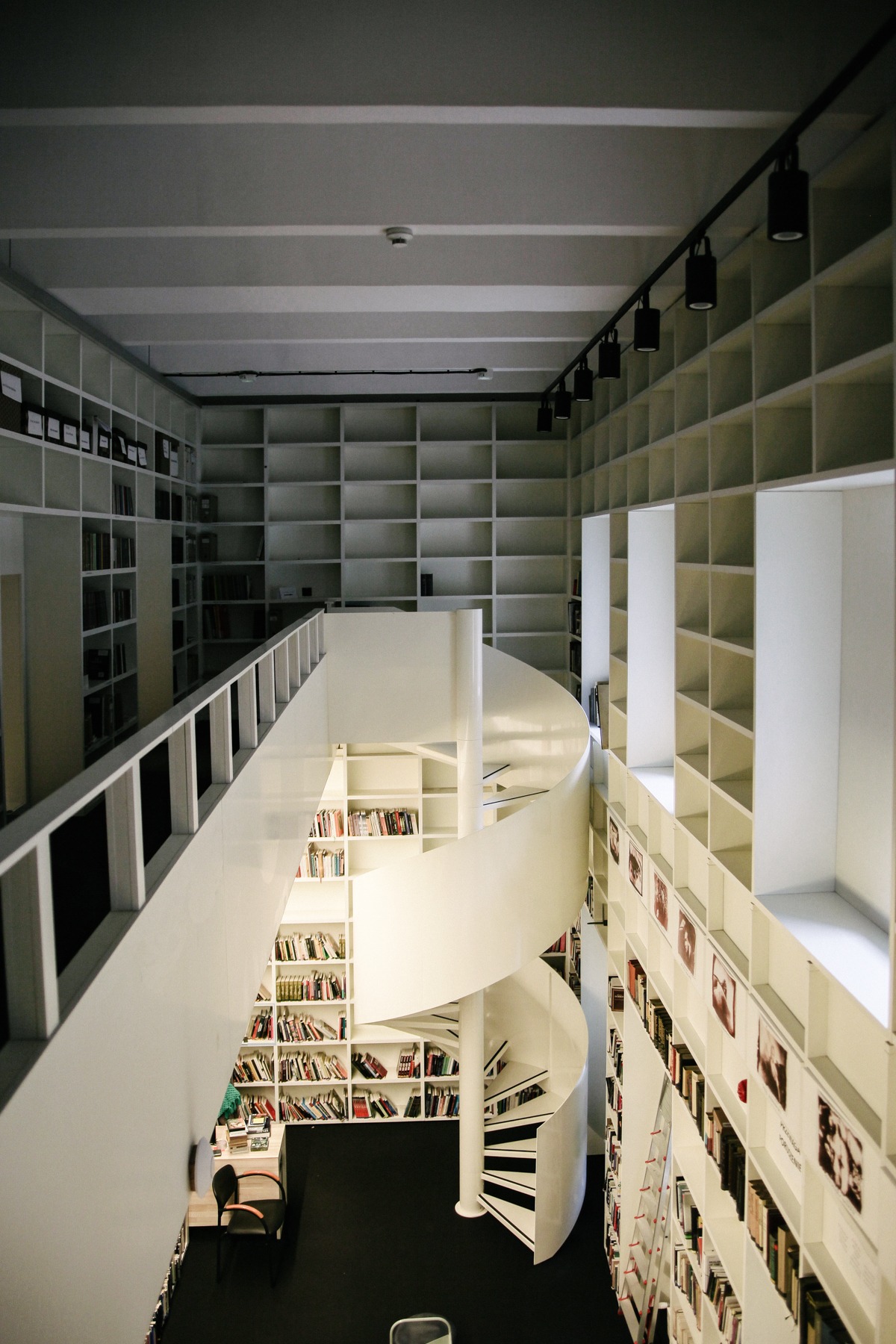 Wnętrze biblioteki z białymi regałami wypełnionymi książkami, spiralne schody na czarnym tle podłogi, oświetlenie szynowe.