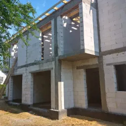 Surowy stan domu z białych bloczków, widoczne drewniane elementy konstrukcji dachu, drabina oparta o ścianę, teren budowy w naturalnym oświetleniu.