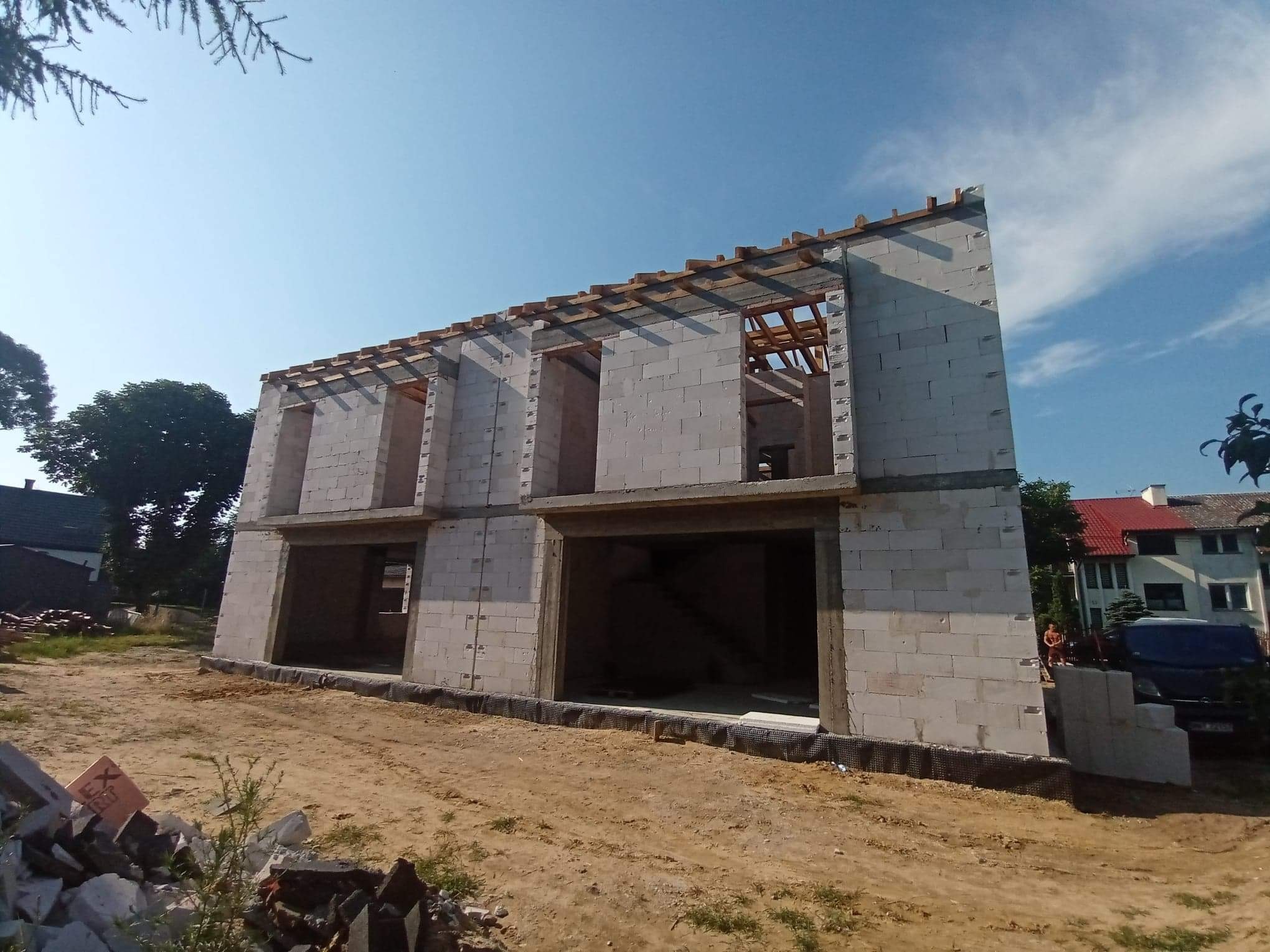 Dwukondygnacyjny dom w surowym stanie, wzniesiony z białych bloczków, z widoczną konstrukcją dachu i otworami okiennymi bez okien, na działce z piaskiem i gruzem.