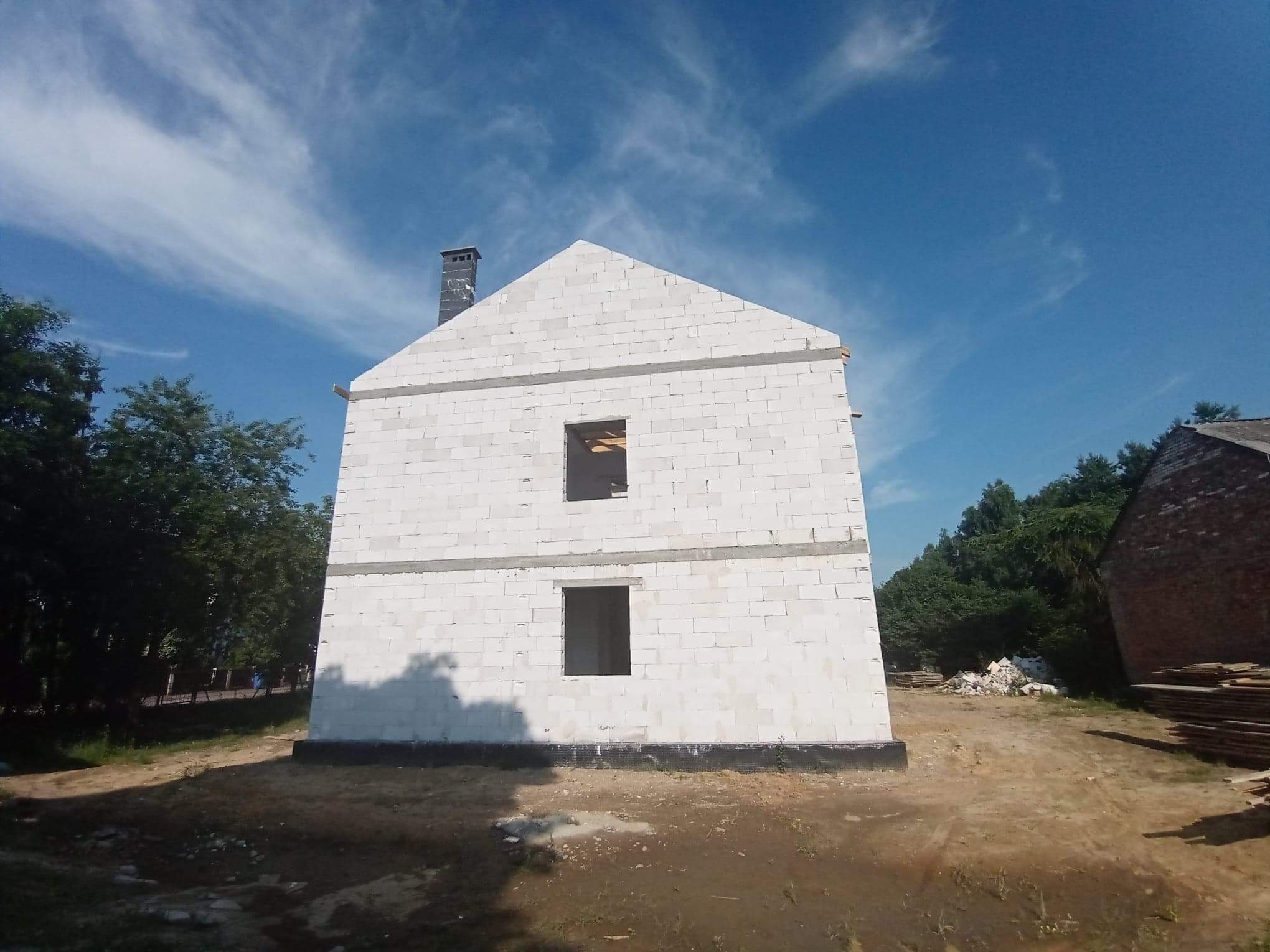 Surowy stan dwukondygnacyjnego domu z białego bloczka, widoczne otwory okienne bez ram, komin z szarej cegły, teren budowy z hałdą gruzu i stertą desek, błękitne niebo z delikatnymi chmurami.