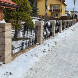 Nowoczesne ogrodzenie panelowe z czarnymi przęsłami i słupkami z jasnego kamienia, widoczne na tle ośnieżonego terenu i żółtego domu z balkonem.