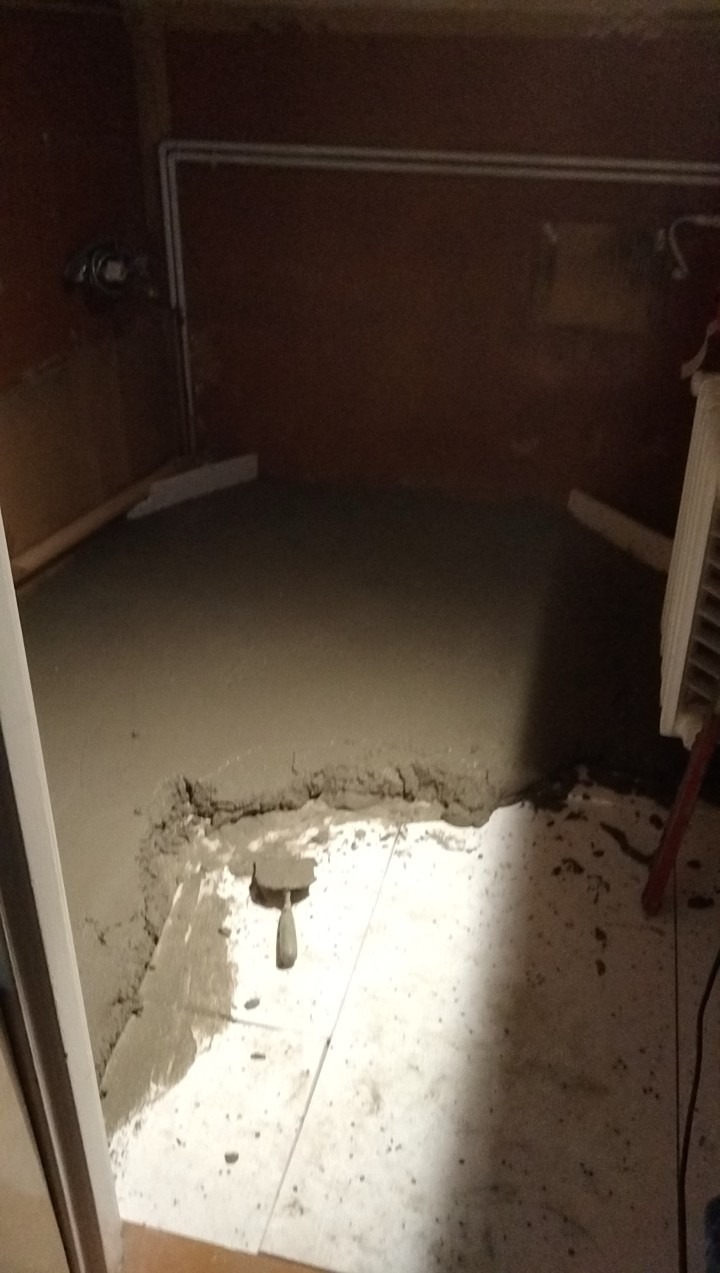 Wylewka podłogowa w trakcie wykonywania, widoczna kielnia na płytkach i grzejnik na ścianie, fragment instalacji wodnej.