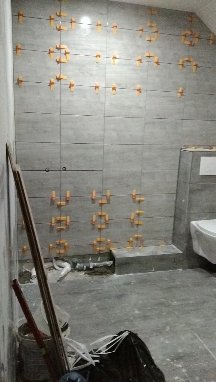 Świeżo ułożone, szare płytki ścienne w łazience, z widocznymi pomarańczowymi krzyżykami dystansowymi, fragment muszli klozetowej w tle, na podłodze widoczne narzędzia i resztki materiałów budowlanych.