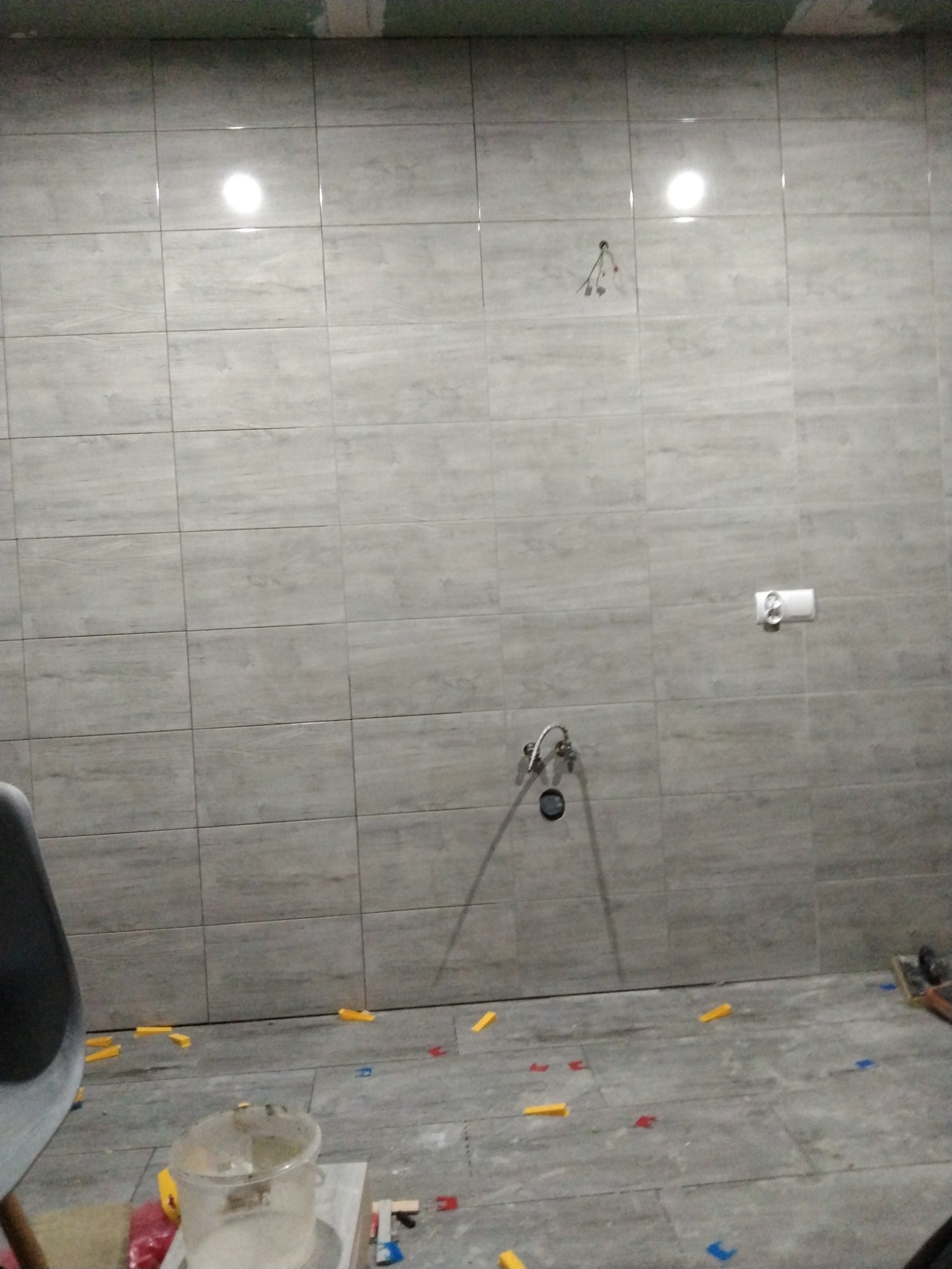 Układanie szarych płytek w łazience, widoczne krzyżyki dystansowe i wiadro z zaprawą, przygotowane do fugowania.