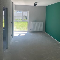 Piotr Warchoł - Wnętrze pokoju po szpachlowaniu i częściowym malowaniu; jedna ściana pomalowana na zielono, pozostałe szare, surowy beton na podłodze, grzejnik przy oknie, butelka wody na parapecie.